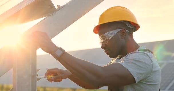 Retrato de cerca del mecánico afroamericano en sombrero duro amarillo trabajando con destornillador en marco de fijación del panel solar. - Metraje, vídeo