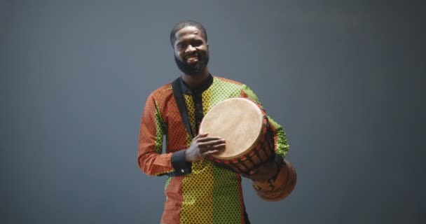 Egy portré fiatal, vidám afro-amerikai férfiról hagyományos ruhában, aki úgy dobol, mintha üdvözölne valakit a partin. Jóképű dobos férfi törzs dob dob mosolyogva boldogan. Zenész játszik Afrikában. - Felvétel, videó