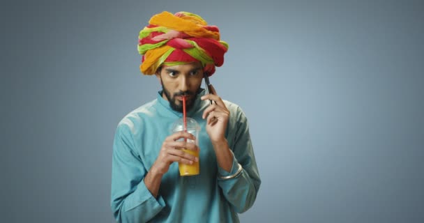 Hindu young handsome man in turban and traditional clothes talking on mobile phone and sipping drink with straw. Indian happy guy speaking joyfully on cellphone and drinking orange juice. - Footage, Video