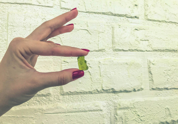 a gummy yellow bear in the hands of a girl with a red manicure. bear are kept on a white brick background. high-calorie homemade dessert made from fruit juices and nectars, a natural product - Photo, Image