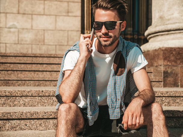 Handsome smiling stylish hipster lambersexual model.Man dressed in white T-shirt.Fashion male posing in the street. Holding, speaking  and chatting in his cellphone. Discussing his good day - Photo, image