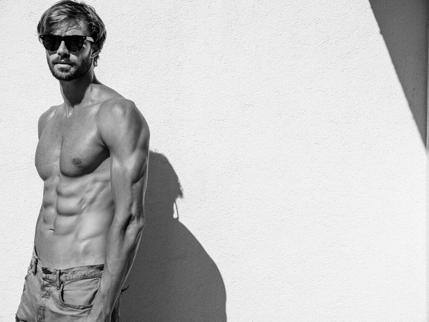 Portrait of sporty handsome strong man. Healthy athletic fitness model posing near white wall in jeans. Confident sexy fashion male with naked nude torso. Lambersexual outdoors in sunglasses - Fotó, kép