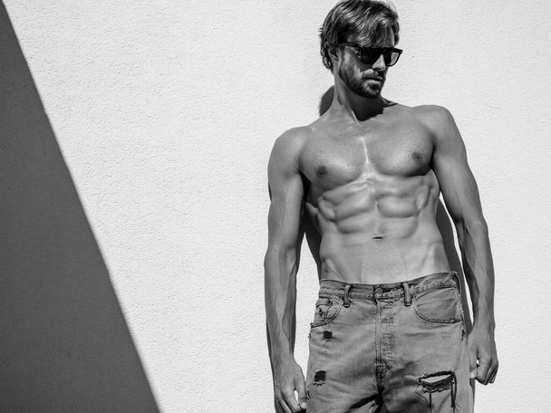 Portrait of sporty handsome strong man. Healthy athletic fitness model posing near white wall in jeans. Confident sexy fashion male with naked nude torso. Lambersexual outdoors in sunglasses - Photo, Image