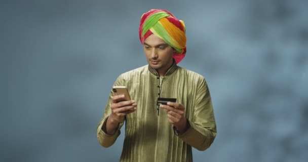 Sarıklı yakışıklı Hindu adam internet üzerinden akıllı telefondan alışveriş yapıyor ve mutlu bir şekilde gülüyor. İnternetten alışveriş yapan ve kredi kartıyla ödeme yapan şık Hintli genç adam. Mutlu erkek alıcı telefonla satın alıyor.. - Video, Çekim