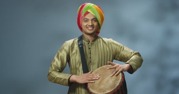 Muotokuva miespuolisesta intialaisesta muusikosta turbaanissa naputtelemassa rumpua ja hymyilemässä iloisesti kameralle. Komea mies perinteisissä vaatteissa leikkii Bongolla iloisella naamalla. Soittimen omaava muusikko. - Materiaali, video