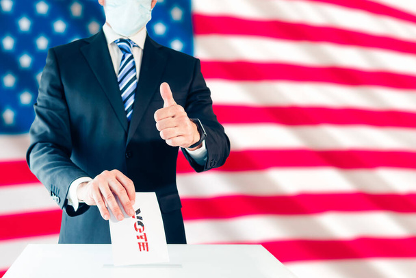 Elegáns férfi védőmaszkban, aki az amerikai elnökre szavaz, és felemeli a hüvelykujját az amerikai zászlóval a háta mögött. Szelektív fókusz - Fotó, kép