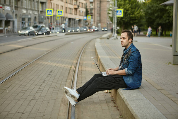 A man with a laptop is waiting for a tramway  at a tram stop. Dangerous behavior on the railway, the male is resting, sitting on the sidewalk near the tracks. - Fotoğraf, Görsel