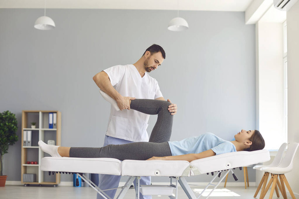 Serious specialist helping young woman with painful knee to do medical exercise and relieve pain - Φωτογραφία, εικόνα
