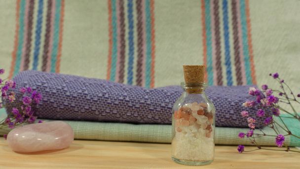 Composição com garrafa de vidro com sais de banho em mesa de madeira com toalhas, pedra rosa, flores roxas e pano com linhas no fundo - Foto, Imagem
