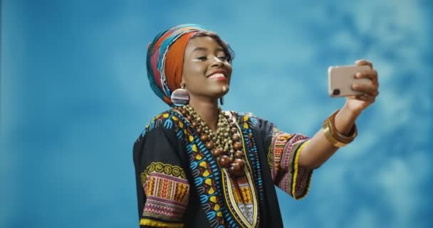 Tyylikäs Afrikkalainen amerikkalainen nuori kaunis nainen perinteisissä vaatteissa hymyilee iloisesti ja poseeraa älypuhelimen kameralle ja ottaa selfiekuvia. Kaunis nainen tekee kuvia puhelimella. - Materiaali, video