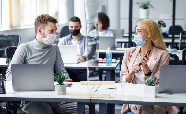Man and woman in protective masks maintain social distance at work. Employees discuss work sitting at table laptop with antiseptic, smartphone and protective glass in interior - 写真・画像