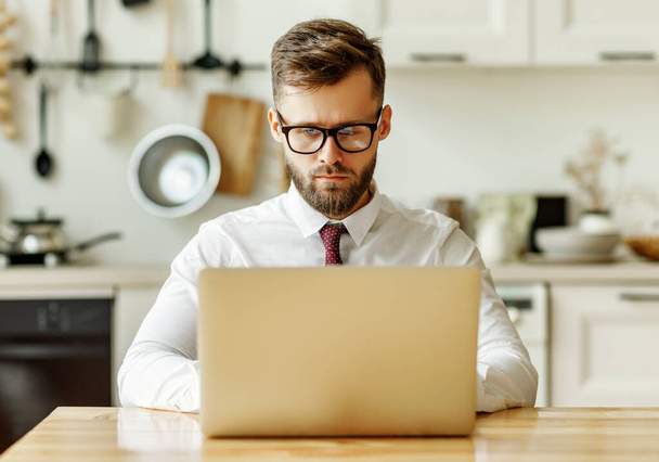 Konzentrierter junger männlicher Unternehmer in festlichem Hemd und Krawatte sitzt am Tisch und benutzt Laptop, während er während der Selbstisolation von zu Hause aus online arbeitet - Foto, Bild