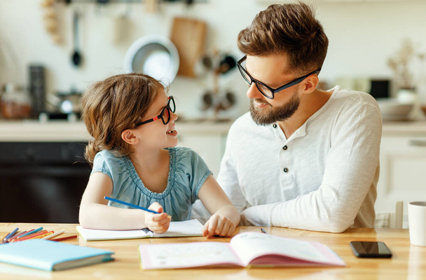 Šťastná rodina: Pozitivní chytrá dívka v brýlích psaní v sešitu a při pohledu na usmívající se otec při domácí úkol společně u stolu v moderních apartmánech - Fotografie, Obrázek