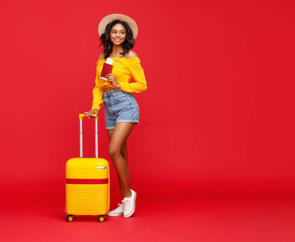 Ganzkörpertrendige Ethnie-Frau mit Pass und Eintrittskarten lächelt in die Kamera und lehnt sich im Sommerurlaub vor rotem Hintergrund an ihr Gepäck. - Foto, Bild