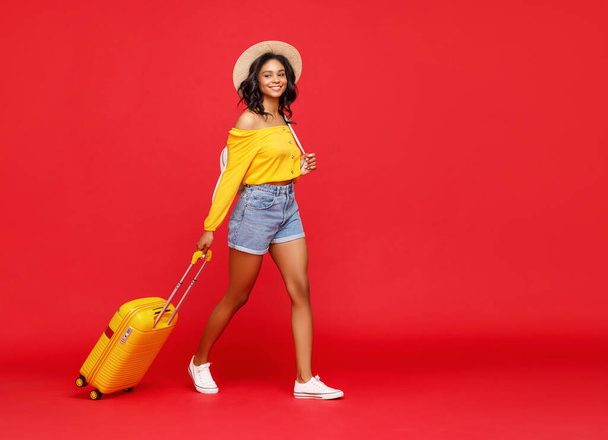 Full length ευτυχισμένη εθνοτική γυναίκα χαμογελώντας, ενώ το περπάτημα στο κόκκινο φόντο και το τράβηγμα luggag - Φωτογραφία, εικόνα