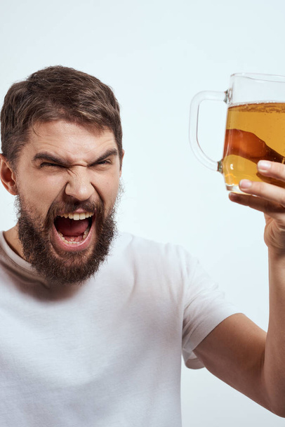 Un uomo barbuto con una tazza di birra su uno sfondo chiaro in una t-shirt bianca vista ritagliata di una bevanda alcolica - Foto, immagini