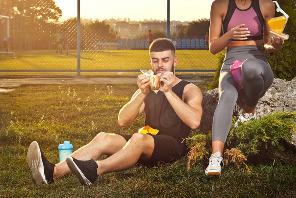 Man and woman eating hamburger after workout. Athletes rest together. Couple on a sunset background. - Фото, изображение