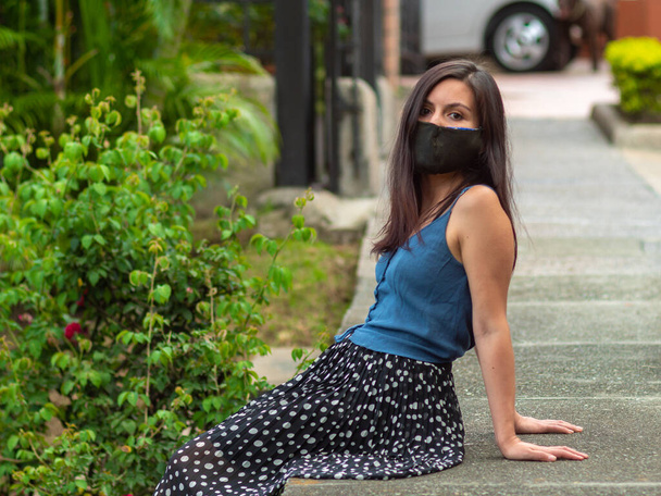 Spaanse jonge vrouw zit op de zijweg met donkere handgemaakte gezicht masker is op zoek naar de camera - Foto, afbeelding