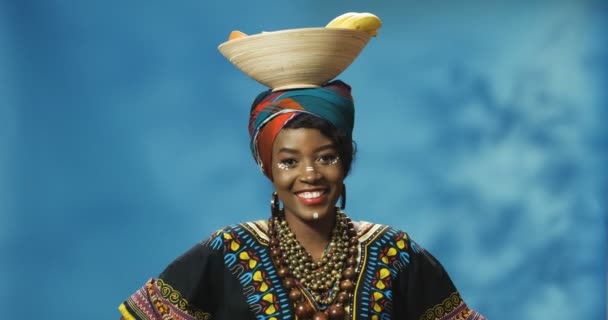 Portret van jonge Afro-Amerikaanse mooie vrouw in tarditionele kleren die vrolijk glimlacht voor de camera en bord met fruit op haar hoofd vasthoudt. Welkom bij Afrika concept. gelukkig meisje poseren met fruit. - Video