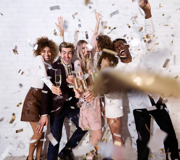 Happy young people celebrating drinking champagne showered with confetti indoor - Photo, Image