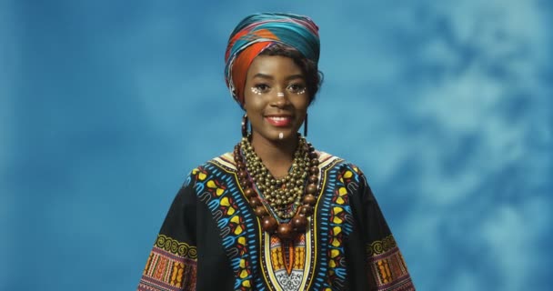 Retrato de una atractiva joven afroamericana en traje tradicional mirando a la cámara con sonrisa. Alegre hermosa hembra en camisa colorida sonriendo felizmente sobre fondo de pared azul. - Metraje, vídeo
