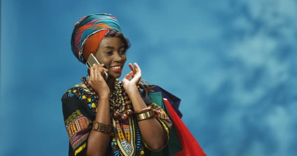Happy young African American woman in turban talking cheerfully on mobile phone, laughing and holding packets over shoulder after shopping. Beautiful girl speaking on cellphone with smile and bags. - Footage, Video