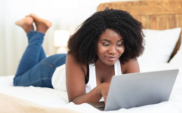 Happy black woman lying on bed, using laptop - Фото, зображення
