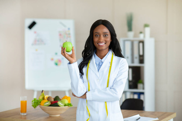 Sonriente asesora de nutrición femenina negra con manzana y cinta métrica mirando la cámara en la clínica - Foto, imagen