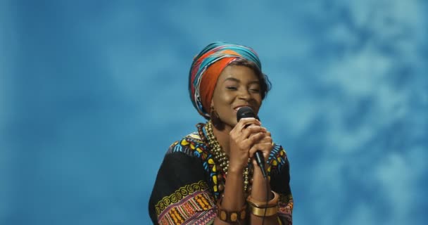 Ευτυχισμένη όμορφη νεαρή κομψή γυναίκα στα παραδοσιακά ρούχα της αφρικανικής φυλής τραγουδώντας σε τραγούδι μικροφώνου και χαμογελώντας στην κάμερα. Αφροαμερικανό χαρούμενο κορίτσι με μικρόφωνο. - Πλάνα, βίντεο
