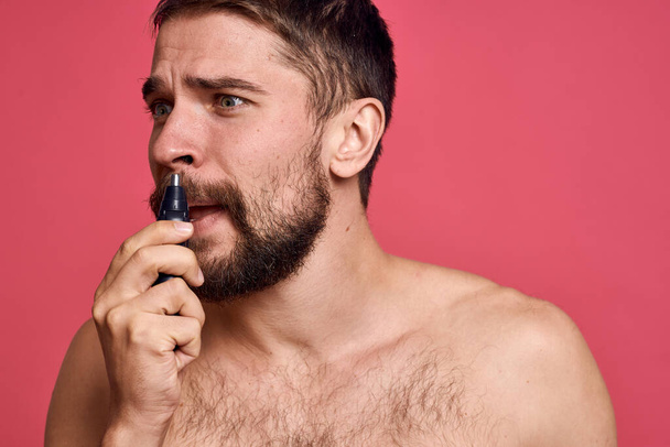 bearded man bare shoulders removes hair from nose body care pink background - Photo, Image