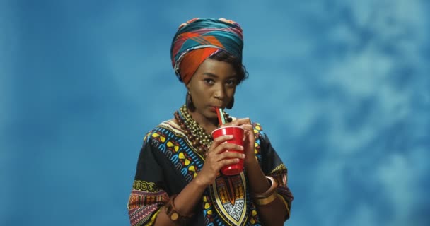 カラフルな服を着た陽気な若い美しいアフリカ系アメリカ人女性の肖像写真は、藁と笑顔でジュースやコークスを飲んでいます。幸せな笑顔魅力的な女の子spping新鮮なドリンク上の青の背景. - 映像、動画