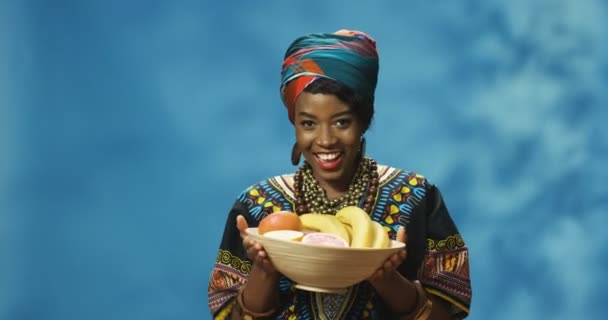 Πορτρέτο του νεαρού χαρούμενα χαμογέλασε όμορφη Αφροαμερικανή γυναίκα χέρι και επίδειξη πιάτο με τροπικά φρούτα και κοιτάζοντας κάμερα. Ελκυστική κοπέλα που δείχνει φρούτα και χαμογελά ευτυχισμένη. - Πλάνα, βίντεο