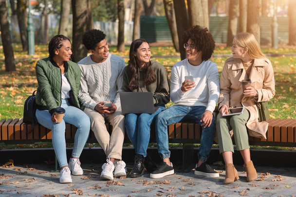 Χαρούμενοι νεαροί φίλοι συζητούν, χρησιμοποιούν gadgets στο πάρκο - Φωτογραφία, εικόνα