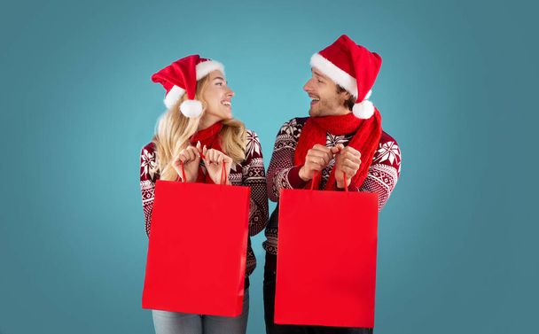 Noel kıyafetleri içindeki genç adam ve kız, mavi arka planda tasarlanmış alışveriş çantalarını gösteriyor. - Fotoğraf, Görsel