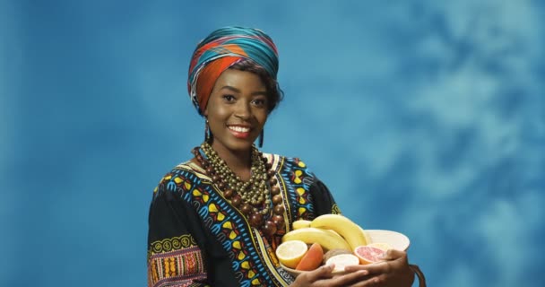 Muotokuva nuoresta iloinen hymyili melko Afrikkalainen amerikkalainen nainen osoittaa lautasella trooppisia hedelmiä ja katsot kameraa. Iloinen kaunis tyttö näyttää hedelmiä ja hymyilee onnellisesti. - Materiaali, video