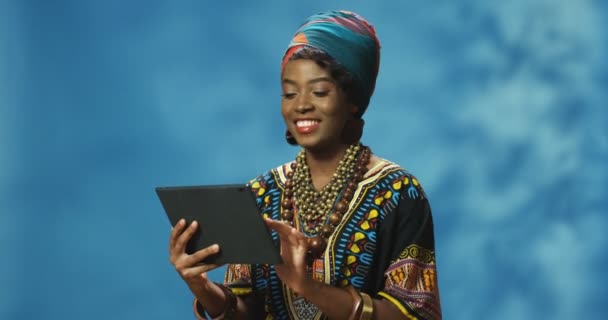 Geleneksel giysili Afro-Amerikan güzel bir kadın elinde tablet cihazı, tıklatıyor ya da kaydırıyor. Tablet bilgisayarda yazan Hintli kızı cezp et. Kadın çevrimiçi video izliyor. - Video, Çekim