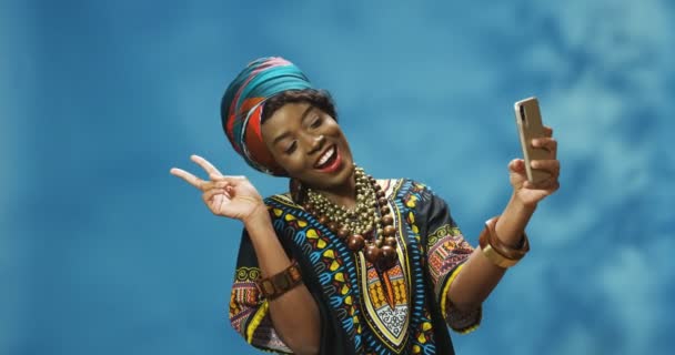 Стильная привлекательная афроамериканская молодая женщина в традиционном наряде радостно улыбается и позирует перед камерой смартфона и фотографирует селфи. Красивая женщина делает фотографии с телефона. - Кадры, видео