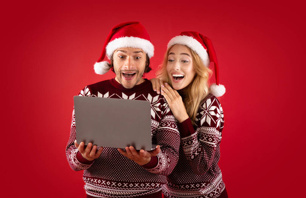 Ενθουσιασμένος νεαρός και η φίλη του κερδίζουν λαχείο Χριστουγέννων σε φορητό υπολογιστή με κόκκινο φόντο - Φωτογραφία, εικόνα