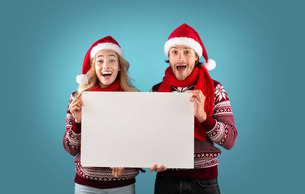 Publicité de Noël. Couple millénaire excité tenant affiche vide avec maquette pour le design sur fond bleu - Photo, image