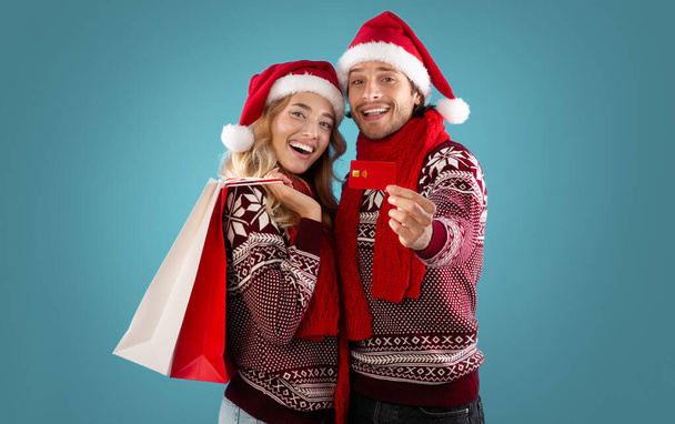 Millennial couple in Santa hats showing credit card and Christmas shopping bags over blue background - Valokuva, kuva