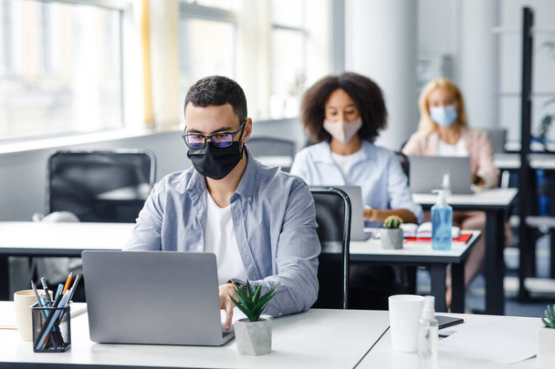 Teamwork in corporate company and returning to work after quarantine covid-19. Focused millennial man in glasses and protective mask works at laptop at workplace with antiseptic - Photo, Image