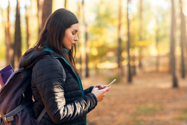 Женщина с рюкзаком с помощью смартфона во время прогулки по лесу - Фото, изображение