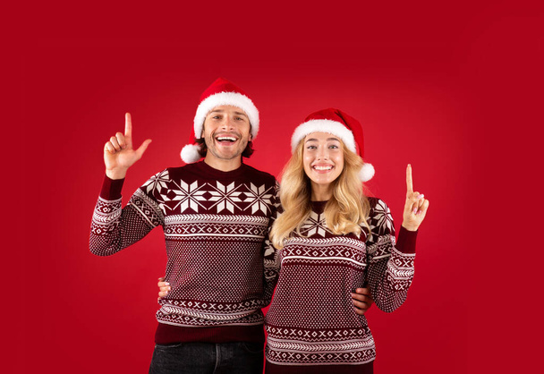 Χαρούμενη κυρία και ο φίλος της με τα χριστουγεννιάτικα πουλόβερ και τα καπέλα του Άη Βασίλη που δείχνουν προς τα πάνω στο κόκκινο φόντο - Φωτογραφία, εικόνα