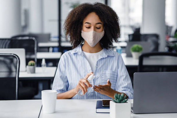Gezondheidsbescherming en nieuw normaal op de werkplek. Afro-Amerikaanse jonge vrouw in beschermende masker desinfecteert handen met antiseptische - Foto, afbeelding