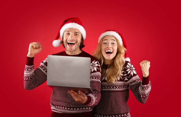 Świąteczna zniżka. Podekscytowany facet i jego dziewczyna z laptopem pokazują gest TAK na czerwonym tle - Zdjęcie, obraz