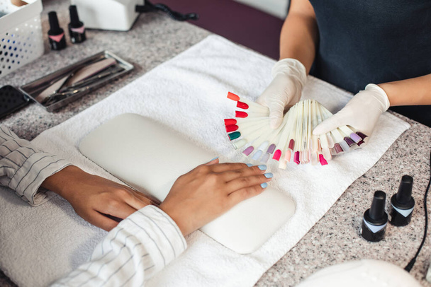 Nail polish color palette. Master in rubber gloves offers samples to african american woman client on table with manicure equipment - Фото, зображення