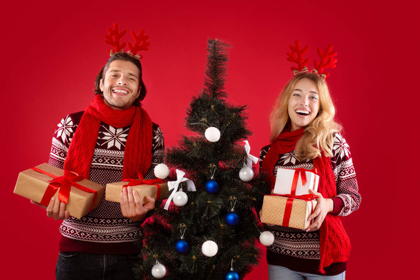 Porträt eines glücklichen jungen Paares mit Weihnachtsbaum und Geschenken auf rotem Hintergrund - Foto, Bild