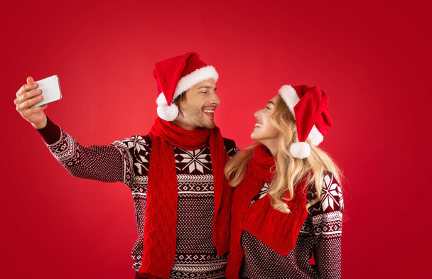 Making memories together. Lovely girl with her boyfriend in Santa hats and sweaters taking selfie over red background - Фото, изображение