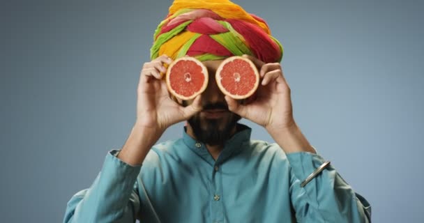 Close up of cheerful Hindu young handsome man in colorful turban holding two halves of grapefruit closing his eyes and opening. Attractive happy guy with fruit in hands portrait in traditional clothes - Footage, Video