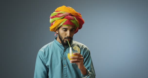 Retrato de un joven y alegre hombre hindú guapo en colorido turbante bebiendo jugo de naranja con paja y sonriente. Feliz sonrió chico atractivo sorbiendo bebida fresca sobre fondo de pared gris. - Metraje, vídeo